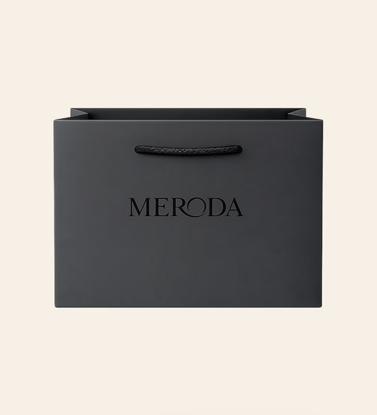 Mattschwarze Geschenktasche mit Kordelgriff und geprägtem Meroda Logo vor cremefarbenem Hintergrund.